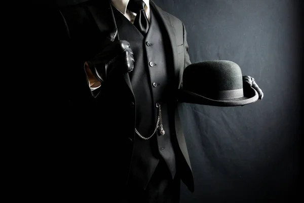 Man in Dark Suit and Leather Gloves Holding Bowler Hat on Black Background. Concept of Classic and Eccentric British Gentleman