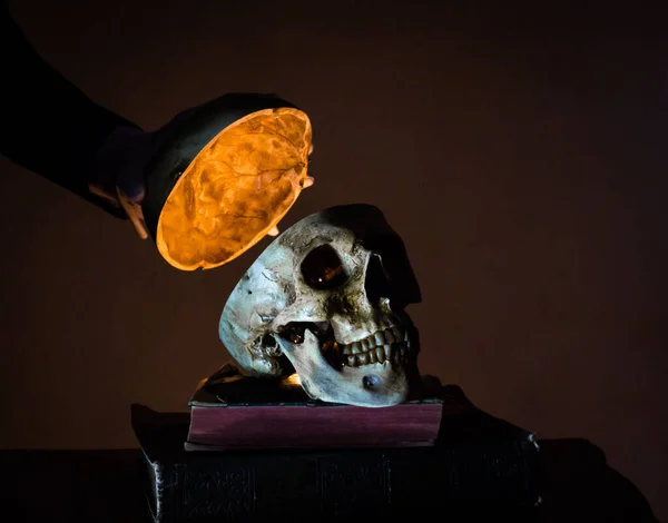 Portrait of Mysterious Figure Lifting the Top of a Human Skull on Stack of Ancient Books. Light After Death. Dark and Haunting.