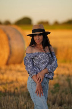 A stylish glamorous model in a straw hat poses in a field against the background of a haystack