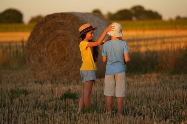 Two children stand in front of a haystack, a girl in a Mexican hat sideways, a boy in a white hat with her back, a girl holding a boy by the head