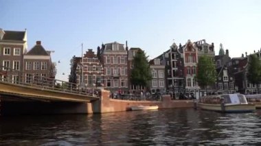 Amsterdam, view of city from river, camera floats under bridge. Traditional Dutch-style urban buildings in summer from river. Amsterdam city skyline sunset timelapse at canal waterfront.