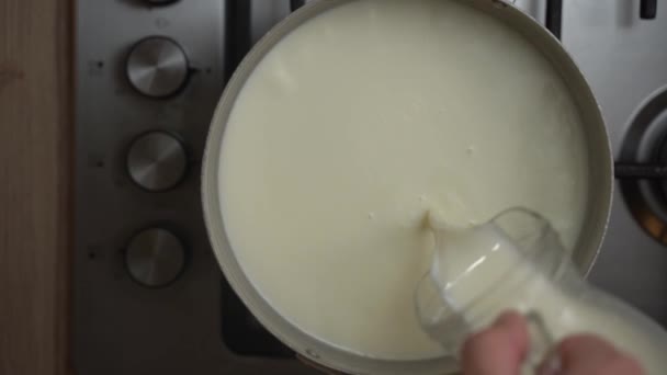 一个人的手从玻璃瓶里把牛奶倒入站在炉子上的铁锅里 满满的牛奶 从上方看去 — 图库视频影像