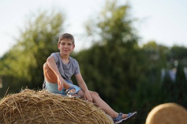 A teenager sits on a haystack on a sunny summer day and smiles at the camera. The guy sits with one leg crossed and his arm hanging down. In the background are haystacks and green trees. Fun childhood
