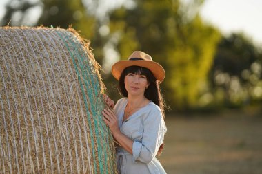A black-haired Ukrainian woman in a straw hat and a light dress peeks out from behind a haystack, a young Ukrainian woman