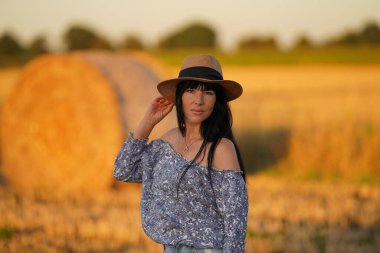 A brunette woman with bare shoulders and long hair holds her hand to a Mexican hat on her head, waist-high view, a girl in a field, behind a haystack and a forest, a healthy diet for healthy skin