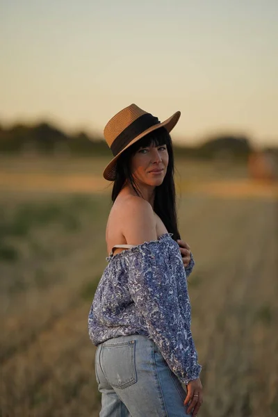 A cowboy woman in a hat and worn jeans stands half a turn on the field and looks into the camera, a country-style woman, a girl in a cowboy hat.