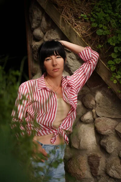 A young Ukrainian woman with black hair leaned against a wall of cobblestones, a view from the bushes. A woman in summer clothes with a bare stomach looks into the camera with her hand behind her head