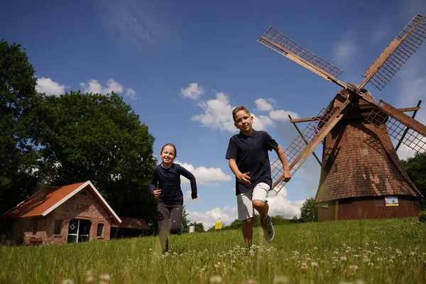 A boy and a girl in summer clothes cheerfully run through a green field, in the background a blue sky, an old building and an old mill. Front view from below. Summer holidays in a German village