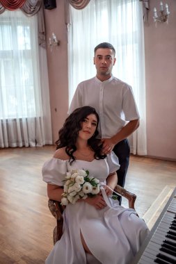 The bride with a bouquet of flowers sits at the piano in the wedding hall, the groom stands behind and looks at the woman he loves. Wedding concept. Beautiful newlyweds
