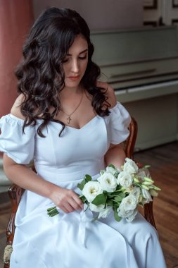 A bride with a bouquet of flowers in a lush white dress sits in a chair and looks into the camera, a portrait of a black-haired Ukrainian bride