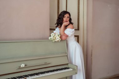 The happy bride in a lush white dress leans on the piano and languidly looks at the bouquet of white roses. Portrait of a happy newlywed