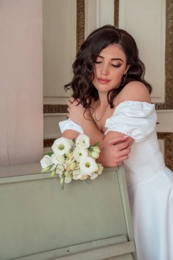 The happy bride in a lush white dress leans on the piano and languidly looks at the bouquet of white roses. Portrait of a happy newlywed.