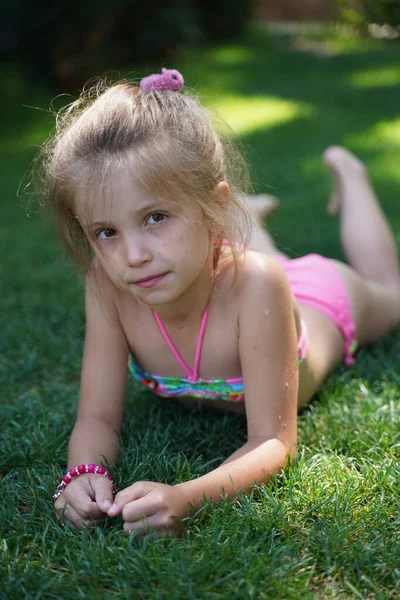 A little blonde girl in a colorful swimsuit lies on the grass and looks into the camera close-up. Summer holidays. Selective focus