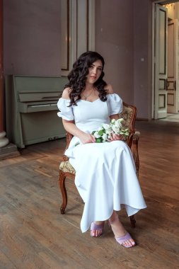 A bride with a bouquet of flowers in a lush white dress sits in a chair and looks into the camera, a portrait of a black-haired Ukrainian bride
