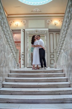 Bride and groom in wedding costumes stand on the stairs in the wedding palace. Beautiful model girl in white dress. Man in suit. Beauty bride with groom. Female and male portrait