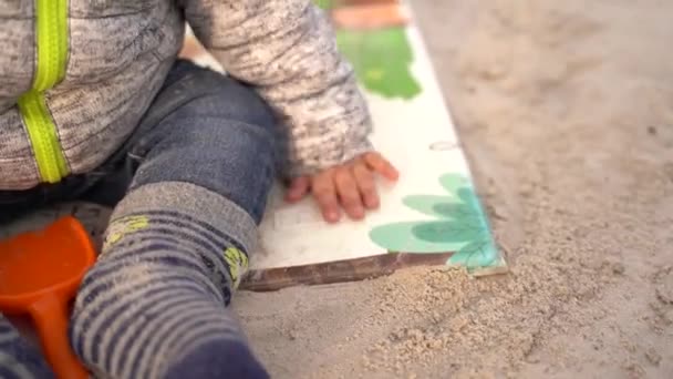 子供は砂の上のマットの上に座り マットを曲げ 砂に沿って彼の手を運ぶ 唯一の腕と体の一部は正面から見える 砂の中で遊ぶ子供 — ストック動画