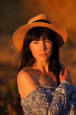A young black-haired girl in a straw hat on a field with a bare shoulder languidly looking into the camera. Attractive model girl on the summer field in a light dress. Portrait of a beautiful girl