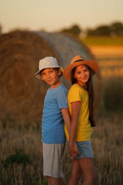 A boy and a girl stand with their backs to each other in bright summer clothes in the field and look smiling into the camera, in the background a haystack and a field.