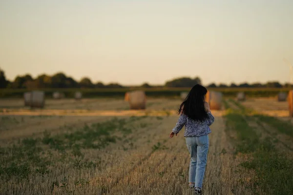 A woman in simple rural clothes and with long black disheveled hair walks through a slanted field from behind view, walking through the field alone, rural boring life