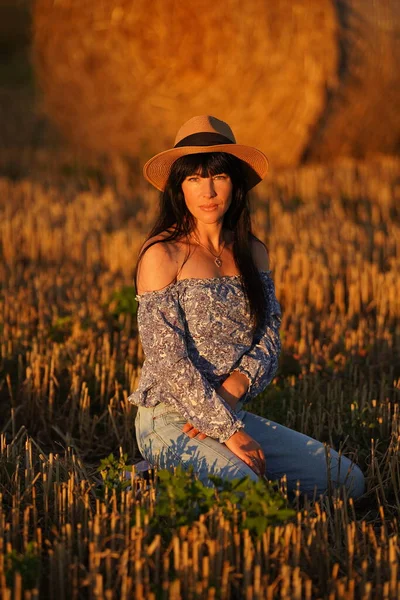 Pretty brunette in a straw hat sits on a mowed field in the summer in rays of the sun and smiles sweetly. Her light blouse is slightly down and you can see sexy bare shoulders covered with long hair