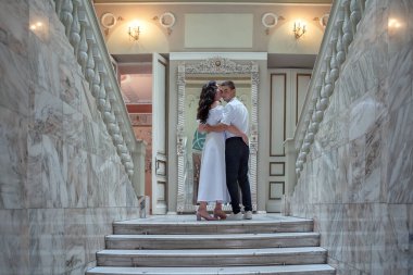 Bride and groom in wedding costumes stand on the stairs in the wedding palace. Beautiful model girl in white dress. Man in suit. Beauty bride with groom.