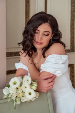 The happy bride in a lush white dress leans on the piano and languidly looks at the bouquet of white roses. Portrait of a happy newlywed. Large portrait