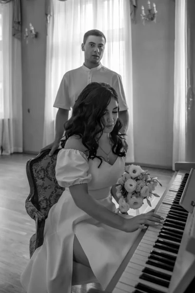 The bride with a bouquet of flowers sits at the piano in the wedding hall, the groom stands behind and looks at the woman he loves. Wedding concept. Black and white photo