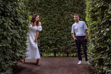 Couple in white clothes have fun in the park in green maze. Bride and groom in white clothes hide in green maze of trees. Newlyweds in summer park among the foliage. Happy young lovers are having fun.