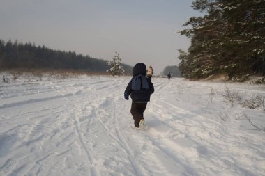 Small child in a winter suit and scarf runs into the distance along a snow-covered road next to the forest, view from behind. A boy running away into the distance on a winter background.