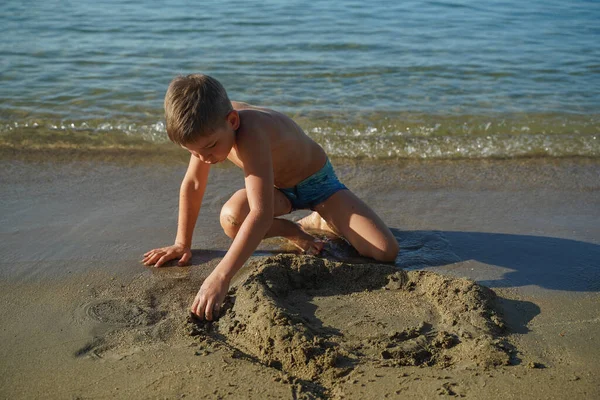 Cute white child builds a sand castle by the sea. Happy boy builds a sand castle on the evening sea beach. A little blond boy with swimming trunks builds a sand castle in the sand on the beach.