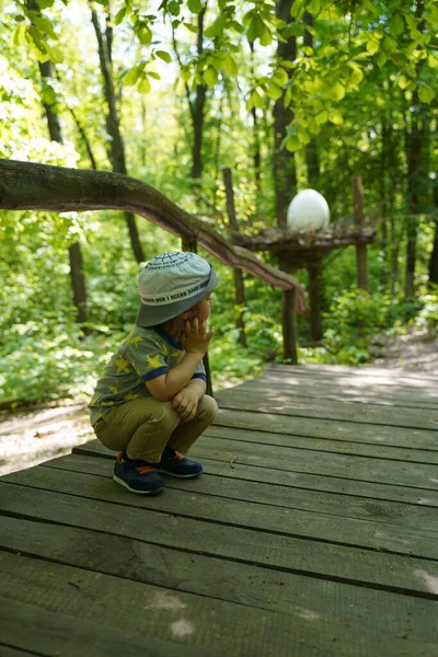 The kid thoughtfully squats in the woods on the bridge and thinks about something turning away from the camera.