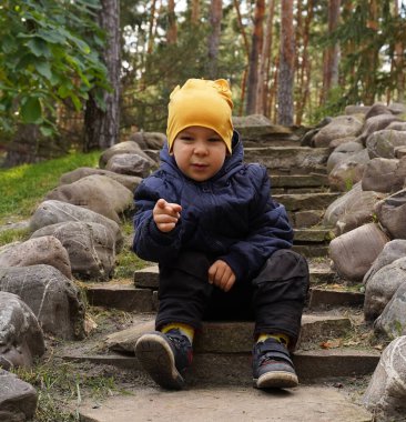 A funny kid in a yellow hat sits on the steps in a landscape park and points his finger at the camera