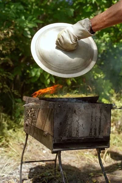 stock image A man opens a lid to check fried potatoes on an open fire. Picnic in the forest. Camping, Lifestyle. Vertical orientation