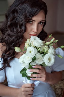A beautiful bride sits on a chair and looks mysteriously at the camera, holding a bouquet of wedding flowers in her hands, an attractive woman in a wedding dress. Happy young woman