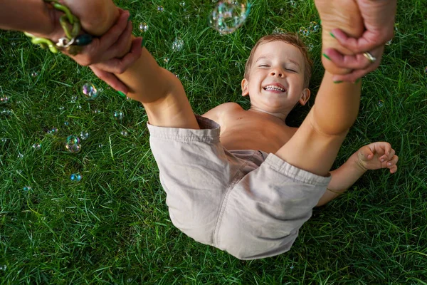 孩子躺在草地上 女人的手托着他的腿 肥皂泡在四处飞舞 一个有趣的夏天生日 软焦点 — 图库照片