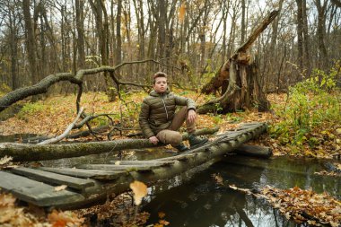 Şık saç stili ve şık giysileri olan genç bir adam köprüdeki ormanda oturuyor. Sonbahar erkek modası.