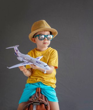 Hasır şapkalı ve yazlık elbiseli küçük bir çocuk elinde oyuncak bir uçakla bir bavulun üzerinde oturuyor. Yaz gezisi. Aile seyahati ve turizm