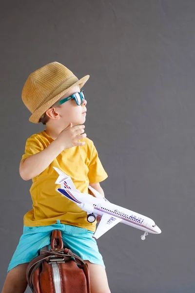 A child in summer clothes and sunglasses sits on a suitcase and holds a toy airplane in his hands. Waiting for travel. Family vacation. Dreams of travel. Summer vacation