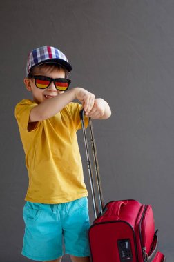 Yazlık elbiseli ve gri arka planda bavulu olan bir çocuk. Stüdyo fotoğrafı. Yaz tatili