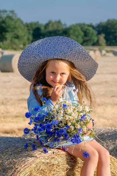 戴着帽子 戴着一束蓝色花朵坐在草堆上的漂亮姑娘 — 图库照片
