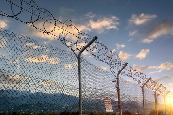A fence made of iron mesh and barbed wire against the backdrop of a beautiful sunset sky and mountains.