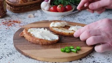 Bir adam krem peynirli ve sebzeli sandviç hazırlar. Domateslerin, soğanların ve yeşilliklerin arka planında.