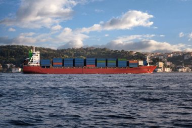 Güvertede konteynırları olan büyük bir nakliye gemisi bulutlu güzel bir gökyüzünün arka planına karşı boğazın yanında yüzer..