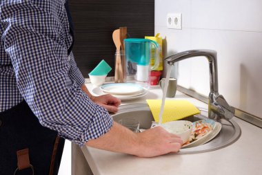 Önlüklü bir adam yemekten sonra kirli bulaşıkları yıkamak üzere. Bir ev mutfağında çalışma alanı..