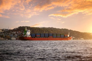 Güvertede konteynırları olan büyük bir nakliye gemisi gün batımındaki gökyüzünün arka planına karşı denizde yüzer..
