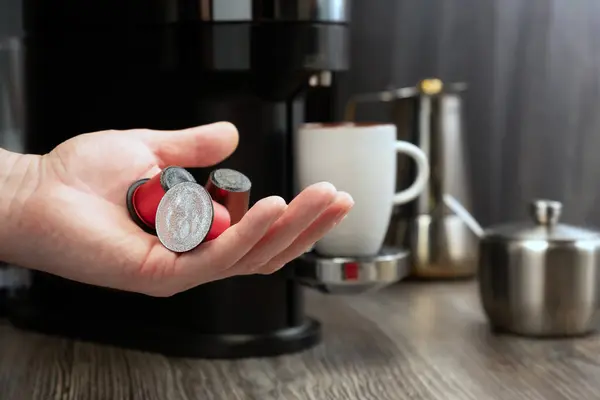 Close-up of a capsule for a coffee machine in a hand against the background of a kitchen with a cup and a coffee machine. Conceptual image.