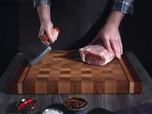 一个男人正要用一把大刀切一大块猪肉肉 在家里的厨房里自制食品烹调概念 免版税图库照片
