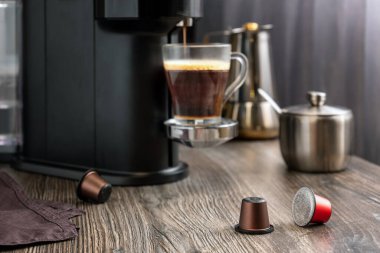 Kahve makinesinde kahve yapmak için kullanılan kapsüller kahve makinesinin arka planında, masanın üzerinde duruyor. Taze aromatik kahve cam bir fincanda hazırlanıyor. Seçici odak.