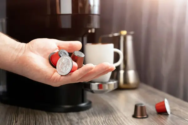 一个人的手拿着装咖啡机的胶囊 放在一个有杯子和咖啡机的厨房的后面 概念形象 图库图片