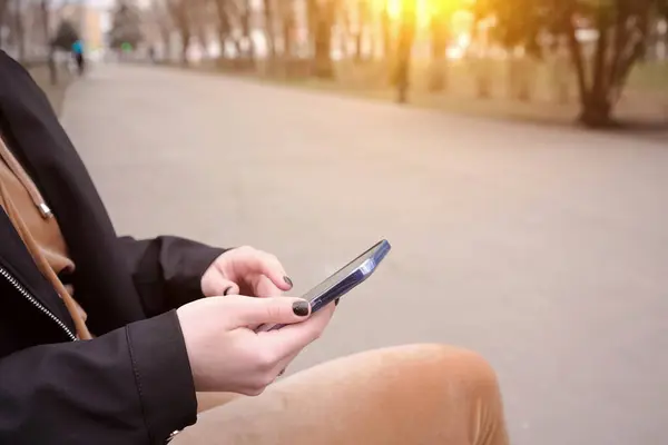 一个女孩手里拿着智能手机坐在城市公园的长椅上 空荡荡的道路 孤独的概念 免版税图库图片
